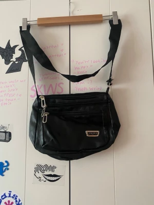 Svart axelväska från vagabond - Snygg svart axelväska från Vagabond i fejkläder med justerbar axelrem. Väskan har flera dragkedjefack framtill och en metallogo på framsidan. Perfekt för att bära det viktigaste och ger en cool vibe till din outfit. Lite sliten på ena sidan av axelbandet.