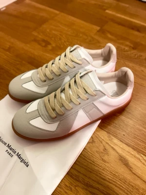 Maison Margiela sneakers - Maison Margiela sneakers i vitt och ljusgrått läder och mocka, med beige skosnören och klassisk gummisula. Snygg retrostil med låg profil och stilrena detaljer. Perfekta för dig som gillar minimalistisk och tidlös design.