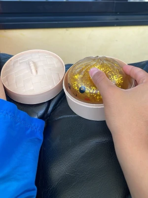 Gul squishy leksak i dim sum-låda - Rolig squishy leksak formad som en gul boll med glitter och ett gulligt ansikte, förpackad i en beige plastlåda som liknar en dim sum-korg. Perfekt att klämma på när man vill slappna av eller leka.
