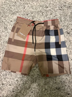 Burberry badshorts med rutigt mönster - Snygga badshorts från Burberry med klassiskt beige, svart, vitt rutmönster. Shortsen har ej blivit använda