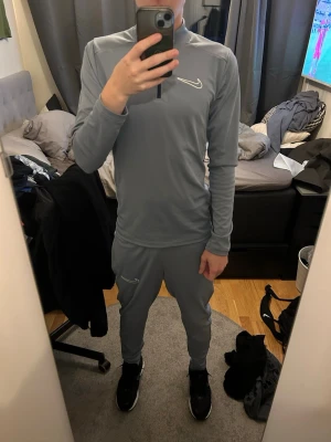  Nike tracksuit herr - Säljer ett grått/ljusblått träningsset från Nike med långärmad tröja och matchande byxor. Setet har vita Nike-loggor på bröstet och benet samt dragkedja vid halsen. Tillverkat i ett mjukt och stretchigt funktionsmaterial som passar perfekt till träning eller chill.