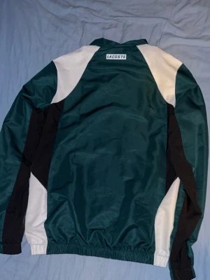 Grön och vit vindjacka från Lacoste - Snygg vindjacka från Lacoste i mörkgrönt med vita och svarta paneler. Jackan har dragkedja framtill, hög krage och elastiska muddar vid ärmar och midja. Perfekt för en sportig look och tillverkad i lätt, vindtätt material.