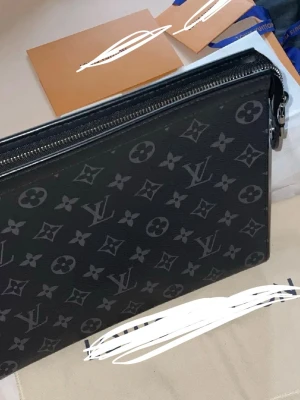 Zara pochette - Snygg svart pochette med klassiskt monogrammönster i grått. Väskan har en rektangulär form, dragkedja upptill och är tillverkad i tåligt läder. Perfekt för att bära det viktigaste med stil.