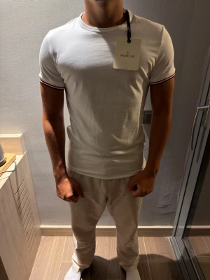 Vit Moncler t-shirt med logga - Snygg vit t-shirt från Moncler med korta ärmar och diskret logga på vänster ärm. T-shirten har ribbade muddar med röd och marinblå rand, vilket ger en sportig vibe. Tillverkad i mjuk bomull och sitter tight för en clean look. Alla storlekar finns