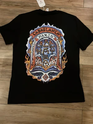 Casablanca - Snygg svart helt oanvänd t-shirt från Casablanca med stort färgglatt grafiskt tryck på ryggen och mindre tryck på bröstet. Klassisk passform och rund hals. Perfekt för dig som vill sticka ut med en unik design. Gjord i mjuk bomull.