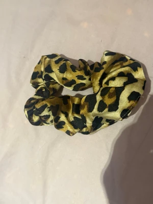 Leopardmönstrad scrunchie - Snygg scrunchie med leopardmönster i guld, svart och bruna toner. Tillverkad i ett glansigt tyg som ger en lyxig känsla och håller håret på plats utan att slita. Perfekt accessoar för att piffa upp din frisyr.