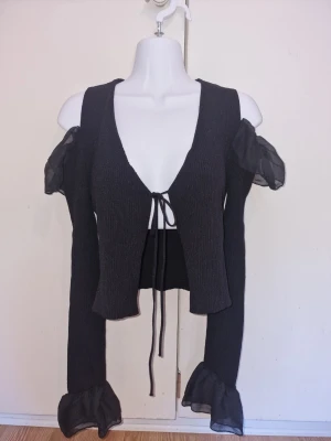 Svart topp med volangärmar från Zara - Svart topp med djup V-ringning och knytband framtill. Toppen har naked shoulders och meshvolanger vid axlar och ärmslut, vilket ger en feminin och unik look. Materialet är ribbat och stretchigt för en snygg passform 🖤