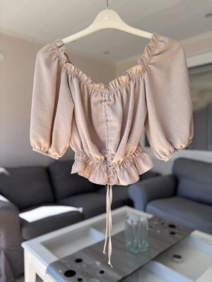 Beige omlott blus med puffärm - Trendig beige blus med omlottkänsla och puffiga trekvartsärmar. Toppen har resår vid axlar och midja, volangkant nedtill och snörning framtill för en snygg passform. 