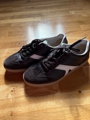 Bruna sneakers med vit detalj, oanvända  - Snygga bruna sneakers med vit sidodetalj och svarta skosnören. Ovandelen är i en mix av mocka och slätt material, vilket ger en stilren och sportig look. Perfekta för dig som vill ha en klassisk men trendig sneaker. 