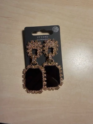 Svarta statementörhängen med strass - Säljer ett par stora, rektangulära örhängen från Monki. De är svarta med gnistrande guldfärgade strass runt kanterna och har en chunky, glamorös look. Perfekta för dig som vill sticka ut och addera lite extra bling till din outfit.