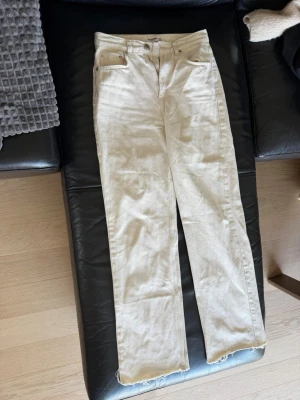 Beige/vit raka jeans med rå kant - Säljer ett par beige/vita jeans med rak passform och rå, fransig kant nedtill. Jeansen har klassisk femficksdesign och knappgylf. Perfekta för dig som gillar en avslappnad och trendig stil.