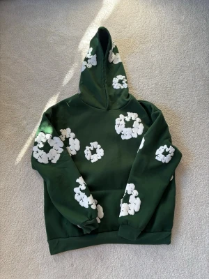 Grön hoodie med vita blommor Denim Tears - Snygg grön hoodie från Denim Tears med unika vita blomapplikationer över hela tröjan. Hoodien har en klassisk huva och ribbade muddar. Perfekt för dig som vill sticka ut med en cool och trendig streetwear-look.