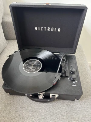 Victrola Journey Portabel Vinylspelare - Snygg portabel vinylspelare från Victrola i svart med inbyggda högtalare, volymkontroll och flera anslutningsmöjligheter som Bluetooth, line in och hörlursuttag. Spelaren har en klassisk resväske-design i hårdplast och metall, perfekt för att spela dina favoritvinylskivor.