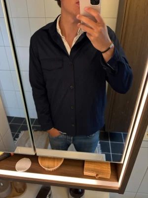 Mörkblå overshirt från Zara - Stilren mörkblå overshirt från Zara med klassisk krage och knappstängning framtill. En bröstficka med lock ger en snygg detalj. Perfekt att bära över en t-shirt eller skjorta för en avslappnad men trendig look.