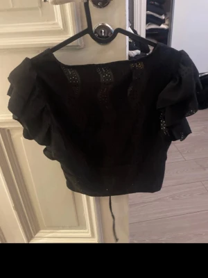 Svart blus med volangärmar och snörning - Svart blus med korta volangärmar och snygga broderade detaljer. Toppen har v-ringning i ryggen och snörning som ger en cool touch. Perfekt för dig som gillar trendiga och feminina plagg med det lilla extra.