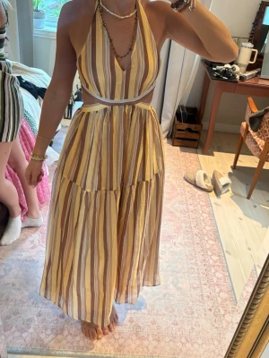 Randig långklänning med öppen rygg - Säljer en somrig långklänning med vertikala ränder i beige, brun och vit. Klänningen har öppen rygg, knyt i nacken och midjan samt djup v-ringning. Tillverkad i ett lätt och luftigt material, perfekt för varma dagar.