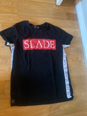 Svart SLÅDE t-shirt med tryck - Svart t-shirt från SLADE med stort rött och vitt tryck framtill och text längs sidorna. T-shirten har korta ärmar och en klassisk passform. Materialet känns som bomull och den har en sportig vibe med tydliga loggor.