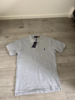 Grå piké Polo Ralph Lauren storlek M - Klassisk grå pikétröja från Polo Ralph Lauren med broderad logga i marinblått på bröstet. Tröjan har korta ärmar, krage och knäppning med två knappar. Tillverkad i mjuk bomull som känns skön mot huden. Kom dm för frågor, bud eller byten 