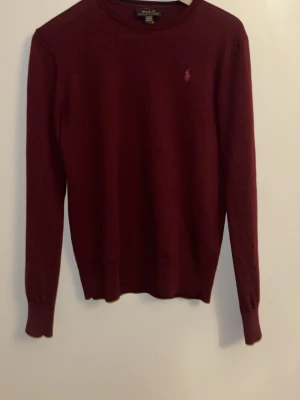 Vinröd stickad tröja Polo Ralph Lauren - Vinröd stickad tröja från Polo Ralph Lauren med rund halsringning och diskret broderad logga på bröstet. Tröjan har långa ärmar och ribbade muddar vid ärmslut och nederkant. Perfekt för dig som gillar klassisk och stilren design. 