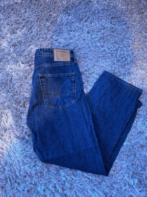 Mörkblå raka jeans från Jack & Jones - Säljer ett par mörkblå jeans från Jack & Jones med klassisk femficksdesign och raka ben. Jeansen har en snygg vintagekänsla och är tillverkade i 100% bomull för en autentisk denimlook. Perfekta för dig som gillar tidlös stil och enkelhet.