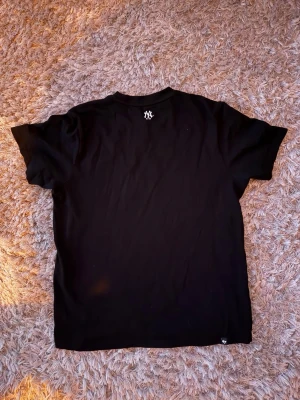 Svart Yankees t-shirt från '47 Brand - Snygg svart t-shirt från '47 Brand med New York Yankees-logga broderad på bröstet och nacken. Klassisk passform med rund hals och korta ärmar. MLB-märke nertill. Perfekt för dig som gillar sportig streetstyle.