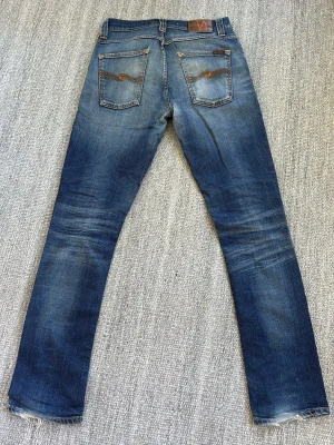Nudie Jeans  - Säljer ett par blå jeans från Nudie Jeans med klassisk orange söm och vågmönster på bakfickorna. Jeansen har normal midja, fem fickor och är tillverkade i denim med snygg slitning och tvättade detaljer.