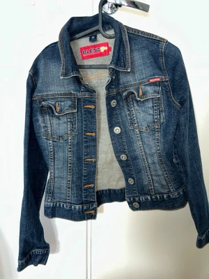 Mörkblå jeansjacka från BlendShe - Snygg mörkblå jeansjacka från BlendShe med klassisk krage, två bröstfickor med lock och knappar samt kontrastsömmar. Jackan har en figurnära passform och silverfärgade knappar framtill. Perfekt för en trendig och avslappnad look.