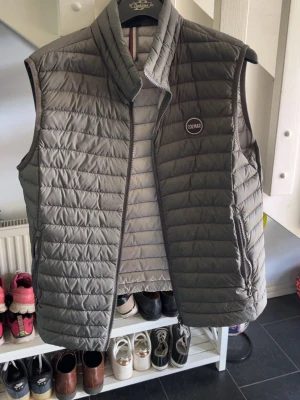 Grå dunväst från Colmar - Snygg grå dunväst från Colmar med quiltad design och hög krage. Västen har dragkedja framtill, två sidofickor med dragkedja och Colmar-logga på bröstet. Perfekt att slänga över en hoodie för extra värme. Materialet är lätt och smidigt.