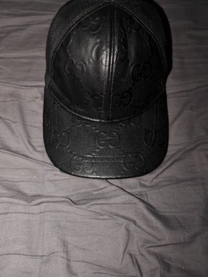 Svart keps från Gucci i läderlook - Snygg svart keps från Gucci med GG-monogram i präglat lädermaterial framtill och mesh baktill för extra ventilation. Justerbar rem bak för perfekt passform. Perfekt accessoar för dig som vill ha en stilren och exklusiv look.