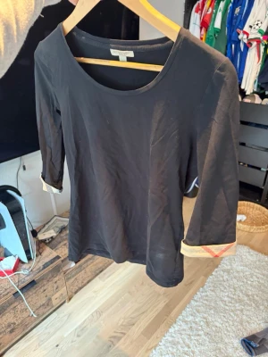 Svart topp från Burberry med trekvartsärm - Stilren svart topp från Burberry med trekvartsärm och rund halsringning. Ärmsluten har den klassiska Burberry-rutiga detaljen på insidan som kan vikas upp för extra stil. Tillverkad i mjuk bomull som känns skön mot huden.