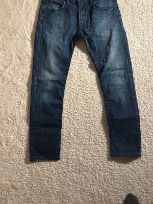 Blå jeans från Lee med raka ben - Säljer ett par klassiska blå jeans från Lee med raka ben och fem fickor. Jeansen har snygga slitningar och en mörkblå tvätt. Tillverkade i slitstarkt denim med Lee-logga på linningen. Perfekta för en avslappnad och trendig stil.