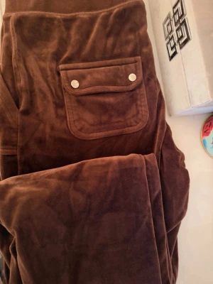Bruna velourbyxor Juicy Couture - Mysiga bruna mjukisbyxor från Juicy Couture i mjuk velour. Byxorna har en ficka bak med tryckknappar och bred resår i midjan. Perfekta för chill dagar och har den klassiska Juicy-looken.