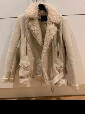 Beige teddyfodrad bikerjacka - Snygg beige jacka från ZARA med mjukt teddyfoder och detaljer som bälte i midjan, dragkedjor och fickor. Jackan har bred krage och fluffiga kanter vid ärmslut och nederkant. 