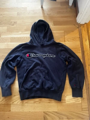 Svart Champion hoodie med broderad logga - Mörk blå hoodie från Champion med stor broderad logga i vitt och rött på bröstet. Klassisk känguruficka framtill och ribbade muddar vid ärmslut och midja. Mjuk bomullsblandning och huva för extra komfort. Perfekt för en avslappnad streetwear-look.