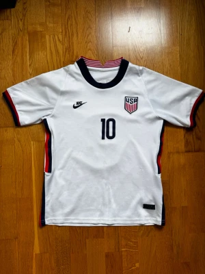 USA Nike fotbollströja Pulisic #10 - Snygg vit fotbollströja från Nike med USA:s landslagsmärke och nummer 10 på bröstet. På ryggen står det Pulisic och nummer 10. Tröjan har marinblå och röda detaljer vid ärmslut och sidor samt randig krage. Perfekt för dig som älskar fotboll och vill stötta USA.