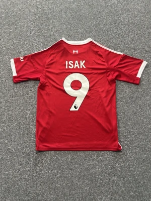 Liverpool matchtröja röd Adidas - Röd Liverpool matchtröja från Adidas med vita detaljer och tryck. Nummer 9 och namnet ISAK på ryggen. Framtill finns Liverpool FC-logga, Adidas-logga och sponsortryck. Tillverkad i lätt och ventilerande material, perfekt för fotboll eller träning.