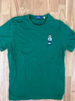 Grön Polo Ralph Lauren t-shirt - Snygg grön t-shirt från Polo Ralph Lauren med klassisk björn-brodyr på bröstet. T-shirten har rund halsringning och är kortärmad. Perfekt för en avslappnad och trendig stil.