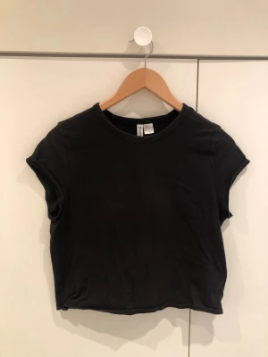 Svart croppad t-shirt från Gina Tricot - En enkel svart croppad t-shirt från Gina Tricot med rund halsringning och korta ärmar. T-shirten har en clean look och är tillverkad i mjuk bomull som känns skön mot huden. Perfekt till jeans eller kjol för en avslappnad stil.