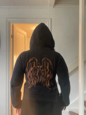 Svart hoodie med änglavingar - Cool svart hoodie med dragkedja och huva. På ryggen finns en hand ritat bronsfärgade änglavingar och på ena ärmen står det ’ANGEL' i samma färg. I huvan står det ’ANGEL’ och hjärtan i samma färg. Hoodien har en tight/ avslappnad passform och är perfekt för en chill y2k look. Tröjan är i storlek XL men jag har sytt den mindre vid midjan. 