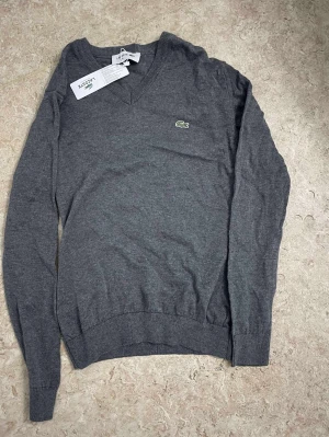 Grå stickad tröja från Lacoste - Klassisk grå stickad tröja från Lacoste med V-ringning och den ikoniska gröna krokodilloggan på bröstet. Tröjan har långa ärmar och ribbade muddar vid ärmslut och nederkant. Perfekt för lager på lager och enkel att matcha.