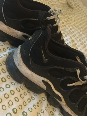 Nike Air Vapormax Plus svarta sneakers - Svarta Nike Air Vapormax Plus sneakers med ikonisk bubblig sula och vita detaljer. Ovandelen är i mesh och syntet med vågiga linjer och små Swoosh-loggor på sidan. Skorna har vita snören och en chunky look som sticker ut.