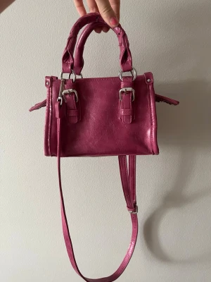 Rosa minihandväska med axelrem - Snygg rosa minihandväska i fejkläder med två flätade handtag, justerbar axelrem och silverfärgade spännen. Väskan har en boxig form och coola detaljer som ger en trendig vibe. Perfekt för dig som vill ha en färgklick till din outfit.        Mått: 14 x 20 x 8 (cm)🤍🤍