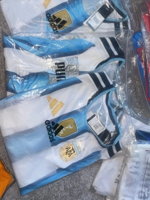 Argentina Adidas fotbollströja - Säljer en officiell Argentina fotbollströja från Adidas med klassiska blå och vita ränder, guldiga detaljer och AFA-emblem på bröstet. Tröjan har svarta Adidas-loggor och rund hals med svarta och vita ränder. Perfekt för match eller träning.