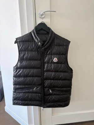 Svart Moncler gui väst - Snygg svart dunväst från Moncler med hög krage och dragkedja framtill. Västen har två fickor med dragkedja och det klassiska Moncler-logotypen på bröstet. Perfekt för lager-på-lager och riktigt stilren look. Moncler påsen medföljer. Priset ej hugget i steg