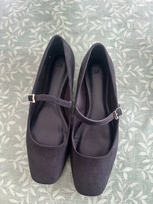Svarta Mary Jane ballerinaskor H&M - Stilrena svarta ballerinaskor från H&M med klassisk Mary Jane-rem och fyrkantig tå. Skorna har en matt finish och justerbart spänne i silverfärg. Perfekta för dig som gillar enkel och trendig design.