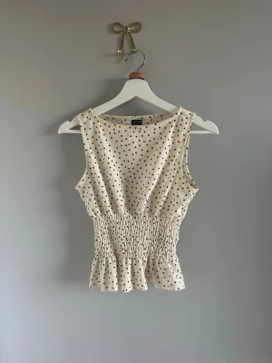 Prickig ärmlös topp med rynkad midja - Snygg ärmlös topp i ljus beige med svarta prickar. Modellen har rynkad midja och volangkant nedtill som ger en feminin siluett. Materialet är mjukt och lätt, perfekt för varma dagar. Passar dig som gillar en trendig och avslappnad look.