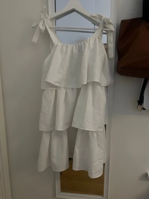 Vit volangklänning från Boohoo - Säljer en vit långklänning från Boohoo med flera volanger och knytband på axlarna. Klänningen har en luftig och flowy passform, perfekt för varma dagar. Tillverkad i bomull för en skön känsla.