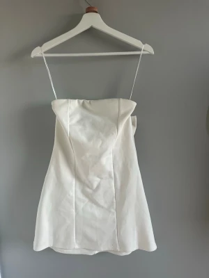Vit kort klänning med rosett - Söt vit kort klänning med tunna axelband och en stor dekorativ rosett i ryggen. Klänningen har en enkel, elegant design och är lätt utsvängd nertill. Perfekt för dig som gillar minimalistisk stil med en twist. Jätte bra kvalitet, tjockt och fint tyg. Inte genomskinligt.