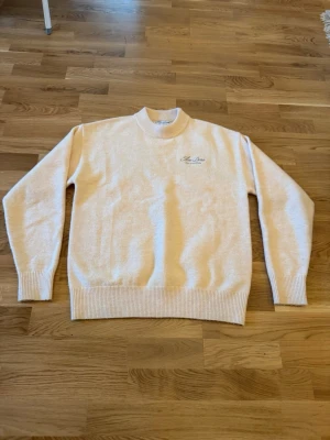 Beige stickad tröja - Beige stickad tröja från Fine Dose med broderad logga på bröstet och stor text på ryggen. Tröjan har ribbade muddar vid ärmslut och nederkant samt en högre rund hals. Perfekt för dig som gillar stilrena och bekväma plagg.
