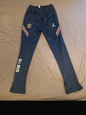 Blå PSG träningsbyxor Jordan - Snygga mörkblå träningsbyxor från Jordan x PSG med vita och röda detaljer på sidorna. Byxorna har PSG-logga, Jumpman-logga och texten PARIS längs benet. Perfekta för fotbollsträning eller chill. (Finns en tröja till på min sida:). 